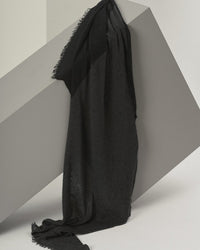 The Grisal Love Duo Cashmere Scarf in Charcoal & Black, featuring frayed edges, is draped over a large slanted gray rectangular block set against a neutral background.