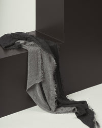 The Grisal Love Duo Cashmere Scarf in Heather Grey & Charcoal, featuring frayed edges, is draped over a sleek dark geometric structure against a minimal white and charcoal background.