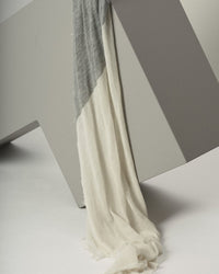 The Grisal Love Duo Cashmere Scarf in Milk & Heather Grey, featuring a two-tone design, drapes over the edge of a geometric light gray structure against a neutral background.