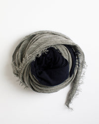 A Grisal Love Duo Scarf in navy and heather grey is rolled up in a circular shape on a white background. The scarf features frayed edges and a soft, textured look reminiscent of luxurious cashmere.
