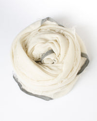 The Grisal Rosa Cascade Cashmere Scarf in Milk X Heather Grey is a soft, light cream-colored scarf with frayed edges and a thin gray border, loosely rolled into a circle on a white background.