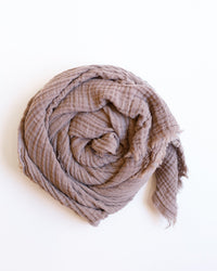 The Freedom Scarf in Lilac by It is well LA is a soft cotton scarf with subtle stripes and frayed edges, rolled up in a circle on a white background for a cozy, relaxed look.