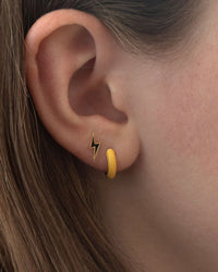 Close-up of an ear with two earrings: a black and gold lightning bolt stud and Kris Nations Enamel Huggie Hoops in Mellow Yellow. Both ear-hugging earrings are crafted in gold vermeil, with brown hair visible beside the ear.