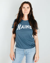 A person with long brown hair wears the Milo in Maine Camp Maine Tee in Dark Blue and light blue jeans, standing against a plain light background.