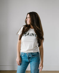 A woman with long brown hair wears the Milo in Maine Camp Maine Tee in Off White and blue jeans, standing against a light wall and looking to her left.