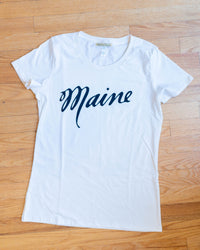 The Milo in Maine Script Maine Short Sleeve Crew in Off White features "Maine" in bold, dark blue script on organic cotton. Laid flat on a light wood floor, it’s perfect for fans of Maine-inspired tees.