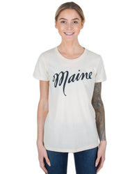 A person with light hair in a ponytail, smiling, wears the Milo in Maine Script Maine Short Sleeve Crew in Off White. They have a tattoo sleeve on their left arm and stand against a white background.