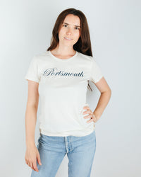 A woman with long brown hair stands against a white background, wearing the Milo in Maine Portsmouth Short Sleeve Crew and light blue jeans. The tee features "Portsmouth" in vintage script. She smiles slightly with one hand on her hip.