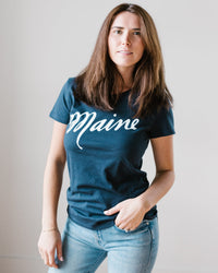 A woman with long brown hair wears the Milo in Maine Script Maine Tee in Dark Blue, paired with light blue jeans. She stands against a plain light background, looking at the camera with a neutral expression.