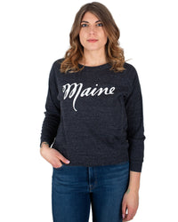 A woman with wavy brown hair wears the Milo in Maine Script Maine Raglan Pullover in dark gray and blue jeans, standing against a plain white background.