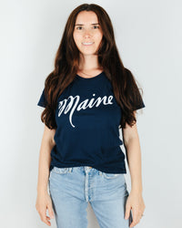 A woman with long brown hair wears the Milo in Maine Script Maine S/S Organic Cotton Tee in Navy, paired with light blue jeans, and stands against a plain white background.