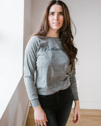A woman with long brown hair stands indoors, wearing the Milo in Maine Script Portsmouth Raglan Pullover in Athletic Gray and dark jeans, looking at the camera with a neutral expression.