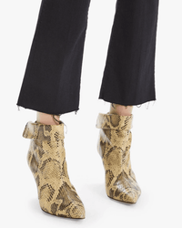 A person stands on a light background wearing Mother’s The Hustler Ankle Fray in Not Guilty, a high rise black denim flare jean with raw hem, paired with pointed-toe yellow and black snakeskin boots.