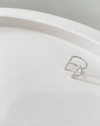 The Nashelle Double Bar Ring in Silver features two parallel bars, elegantly arranged on a smooth white round surface under soft lighting.
