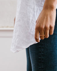 A person wearing a white shirt and dark jeans stands with their arm relaxed, showcasing the Nashelle Faceted Bar Ring in Gold on their finger against a softly blurred background.