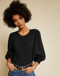 A person with short, curly hair wears the Nation LTD Loren Smocked Peasant Tee Reverse Slub in Jet Black, styled with puffed sleeves and blue jeans, posing hands-in-pockets against a plain beige background.