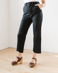 A person stands indoors against light walls and a wood floor, wearing Prairie Underground’s Denim Boiler Pant in Black—a high-rise, cropped flare—paired with brown platform sandals. Only their lower body is visible, one hand in the pocket.