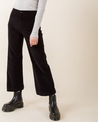 Wearing Prairie Underground’s Knack Cords in Black, a light gray long-sleeve top, and black ankle boots, the person stands against a beige background, shown from shoulders down with one arm relaxed.