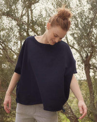 A woman with red hair in a bun smiles softly outdoors among trees, her Frank & Eileen Olive Capelet in British Royal Navy adding an effortless touch to her look as she wears a loose black shirt and light gray pants.