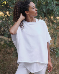 A woman with curly hair in a ponytail stands outdoors among lush green trees, wearing the Frank & Eileen Olive Capelet in White and white pants. She looks to the side, touching her hair, surrounded by natural scenery.