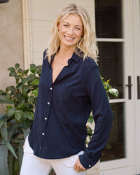 A smiling woman with wavy blonde hair stands outdoors, wearing the Frank & Eileen Eileen Knit Button Up in British Royal Navy and white pants, one hand casually in her pocket, against a backdrop of plants and a beige wall.