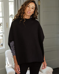 A woman with wavy brown hair wears the Frank & Eileen Audrey Funnel Neck Capelet in Black over a patterned long-sleeve shirt, standing indoors by a white chair and gray paneled wall, looking slightly toward the camera.