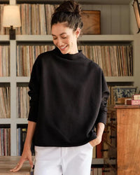 A woman with dark hair in a bun smiles as she looks down indoors, wearing the Frank & Eileen Effie Capelet in Black and white pants. Behind her is a bookshelf filled with vinyl records and a wooden drawer.