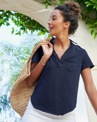 A woman with curly hair in a bun wears the Frank & Eileen Charlotte Perfect Polo in British Royal Navy and white pants. She carries a large woven bag, standing outdoors under a leafy arch, smiling as she looks to the side.