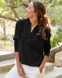 A woman with long brown hair in a ponytail wears the Frank & Eileen Patrick Henley in Black and white pants, standing outdoors among greenery and sunlight, smiling and looking to the side.