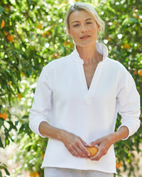 A woman with light hair pulled back stands outdoors before an orange tree, calmly looking at the camera while holding an orange and wearing the Frank & Eileen Patrick Henley in White Terry.