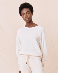 A woman with short curly hair wears the Great’s College Sweatshirt in Washed White, paired with matching drawstring pants, standing against a beige background and looking directly at the camera with a neutral expression.