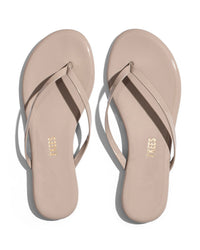 The Tkees Glosses Flip Flop in Custard features a beige minimalist design with thin straps and gold Tkees branding on the insoles, displayed from above on a white background.