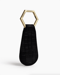The Drop in Black & Gold Basket Weave by TopTote is a black leather woven keychain with a hexagonal gold ring, engraved with “ALBANESE” and “TUESDAY,” perfect as a chic handbag accessory, shown on a white background.