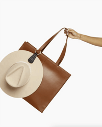 A hand holds The Drop in Black & Gold Basket Weave tote by TopTote, with a beige wide-brimmed hat hanging from one handle using a stylish TopTote hat holder, all set against a plain white background.