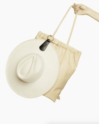 A hand holds TopTote’s The Drop in Black/Gold tote bag with a wide-brimmed cream hat attached by a magnetic hat clip to the handle, set against a plain white background.