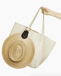 A hand holds The Fray in Black & Gold tote by TopTote, a large cream-colored bag with long straps and a straw sunhat attached using a magnetic hat clip. The background is plain white.