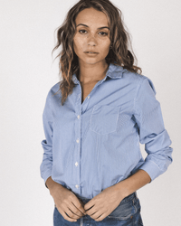 A woman with wavy brown hair and freckles stands against a plain background, wearing the Trovata Birds of Paradis Grace Classic Shirt in Blue/White Stripe with jeans, looking directly at the camera with a neutral expression.