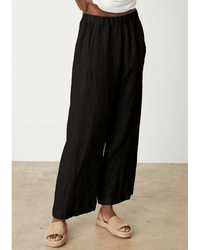 Person wearing Velvet by Graham & Spencer's Lola Pull On Pant in Black, paired with a white top and beige espadrille sandals, standing on a light concrete floor against a plain white wall.