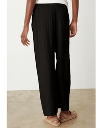A person stands indoors on a beige floor, wearing Velvet by Graham & Spencer's Lola Pull On Pant in Black with a white top and tan sandals. Only the lower half of their body is visible from the back.