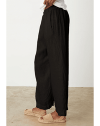 A person stands sideways wearing the Lola Pull On Pant in Black by Velvet by Graham & Spencer, paired with a white top and beige espadrille sandals, on a light tan floor with a plain white wall background.