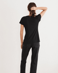 A person stands with their back to the camera, wearing the Velvet by Graham & Spencer Odelia Scoop Neck Tee in Black and dark jeans, one hand holding their hair against a plain white background.