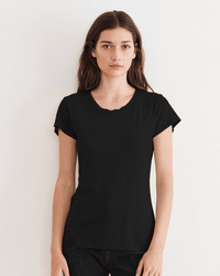 A woman with long brown hair stands against a plain light wall, wearing the Velvet by Graham & Spencer Odelia Scoop Neck Tee in Black and dark pants, looking directly at the camera with a neutral expression.