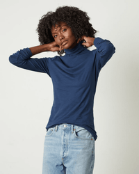 A person with curly hair wears the Velvet by Graham & Spencer Talisia Turtleneck Top in Midnight and light blue jeans, standing against a plain light background with their hands touching the back of their neck.
