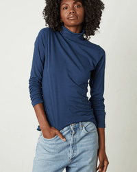 A person wearing the Talisia Turtleneck Top in Midnight by Velvet by Graham & Spencer and light blue jeans stands against a plain background with one hand in their pocket, looking confidently at the camera.