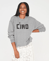 A smiling woman with long braids wears the Clare V. Block Ciao Oversized Sweatshirt in Grey with Black and a white lace skirt, standing against a plain light background.