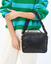 A person in a green and blue striped sweater and white pants carries a Clare V. bag featuring the Braided Leather Shoulder Strap in Black Italian Nappa.