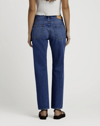 Someone is shown from the back wearing Edwin’s Simone in Idle indigo straight leg midrise jeans, a white sleeveless top, and black sandals, standing against a plain light background.