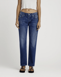 Wearing Edwin's Simone in Idle indigo wash midrise straight leg jeans, paired with a white sleeveless crop top and black sandals, this person stands against a plain light background.