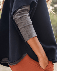A person stands outside with a hand in their pocket, wearing a gray long-sleeve shirt layered beneath the Frank & Eileen Audrey Funnel Neck Capelet in British Royal Navy. Only their torso and arm are visible.