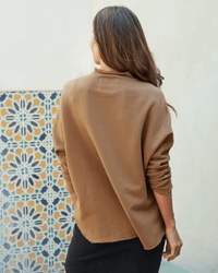 A woman with long brown hair, wearing the Frank & Eileen Effie-L/S Funnel Neck Capelet in Camel and a black skirt, stands with her back to the camera near a wall adorned with colorful geometric tile designs.